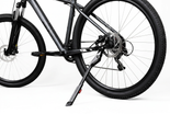 Close-up of a dark gray mountain bike with its kickstand deployed, showing the rear wheel, disc brake, chainring, and frame against a clean white background. 4