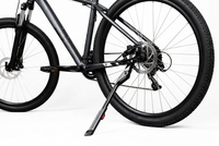 Close-up of a dark gray mountain bike with its kickstand deployed, showing the rear wheel, disc brake, chainring, and frame against a clean white background. 4