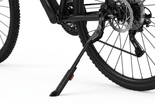 Close-up of a bicycle's rear wheel and black kickstand on a clean white background, showing the stand supporting the frame near the wheel hub. 1